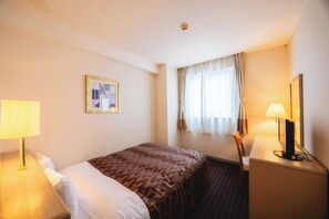 A hotel room with a bed, a desk with a computer, a lamp, and a window with curtains.