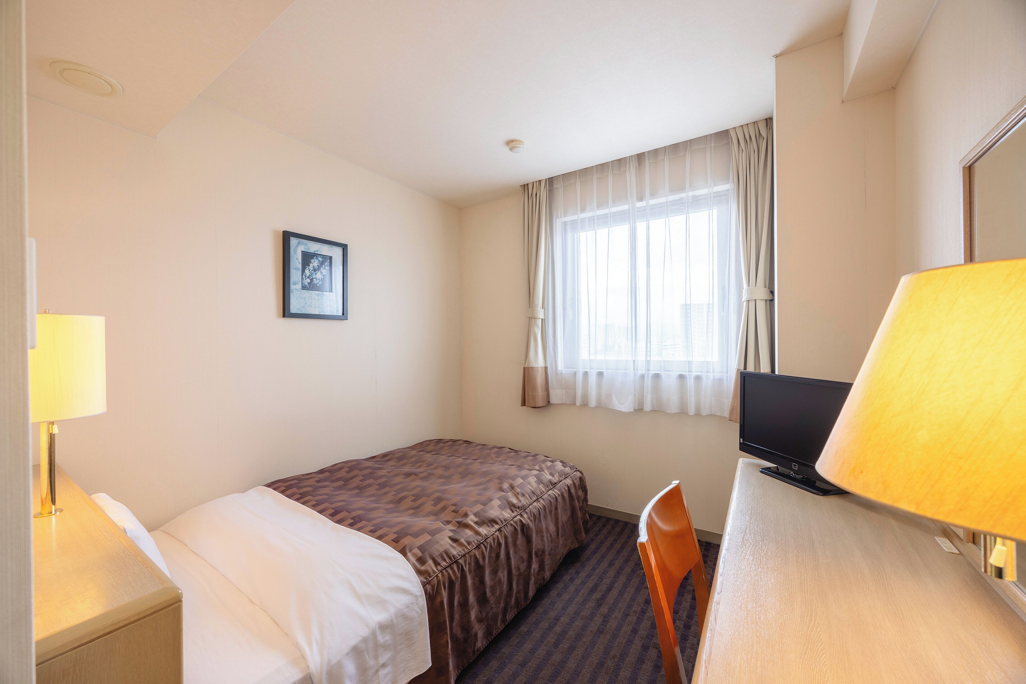 Ein Hotelzimmer mit einem Einzelbett, einem Schreibtisch mit Computer, einem Fenster mit Vorhängen und einem gerahmten Bild an der Wand.