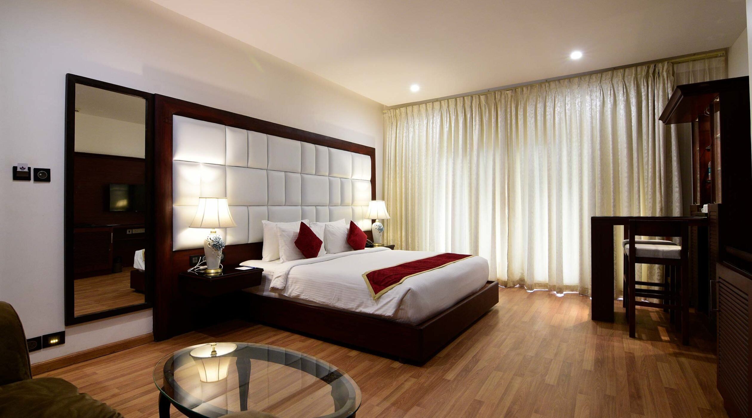 A hotel room with a large bed, a bedside table with lamps, a round glass table, a mirror, and a TV.