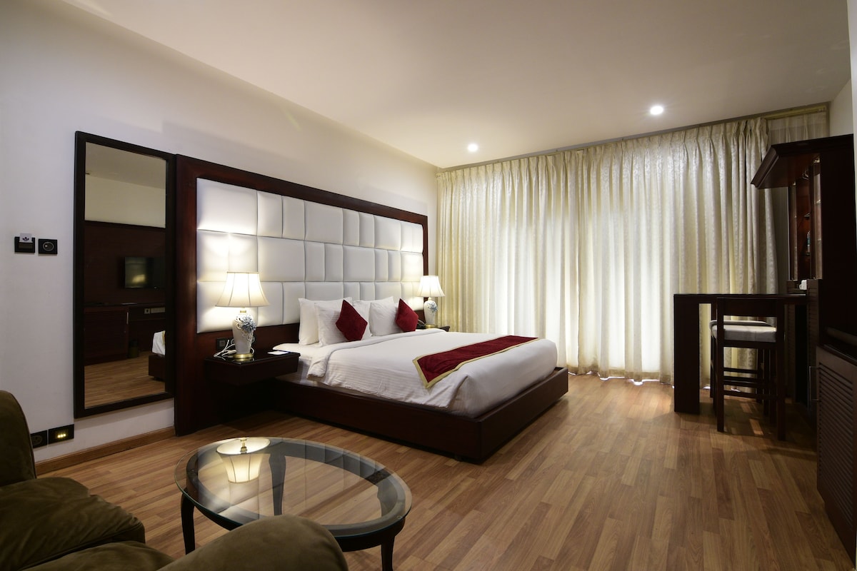 Quarto de hotel com uma cama grande, mesa de cabeceira com luminárias, mesa redonda de vidro e um espelho.