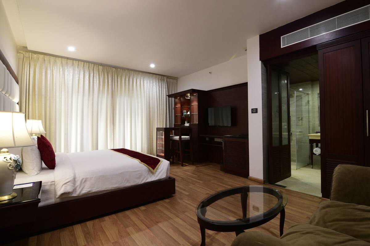 A hotel room with a bed, a desk, a chair, a TV, and a glass door leading to a bathroom.
