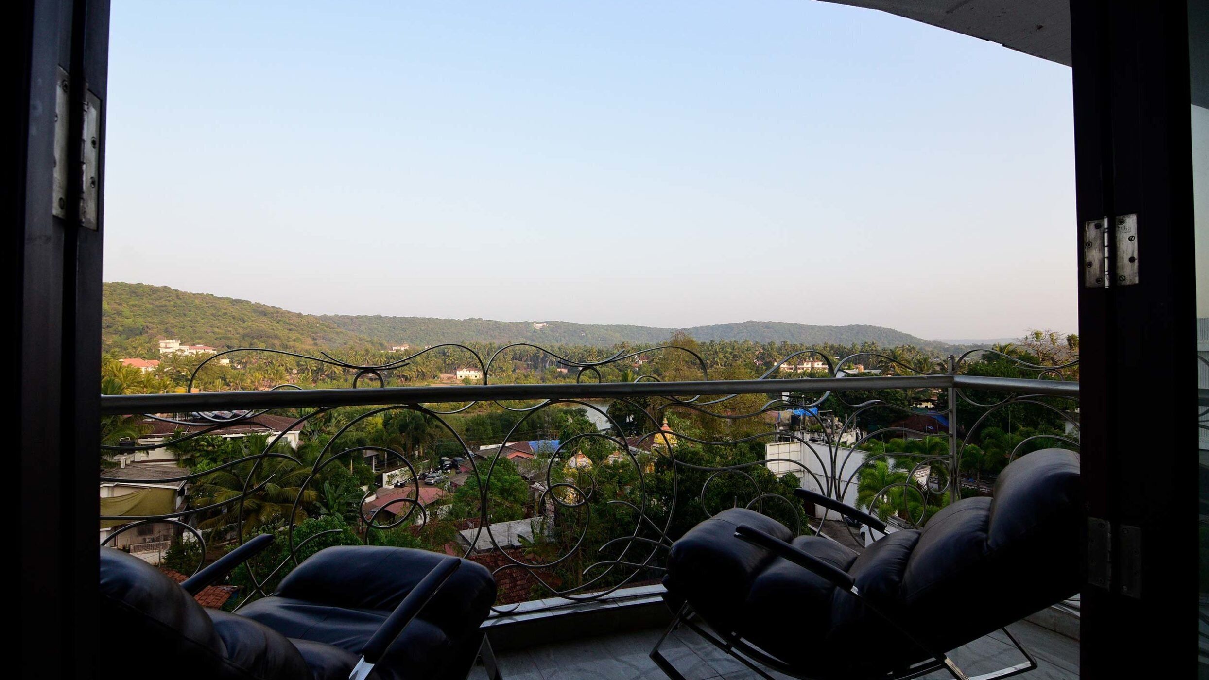 A balcony with two black chairs and a view of a hilly landscape with buildings.