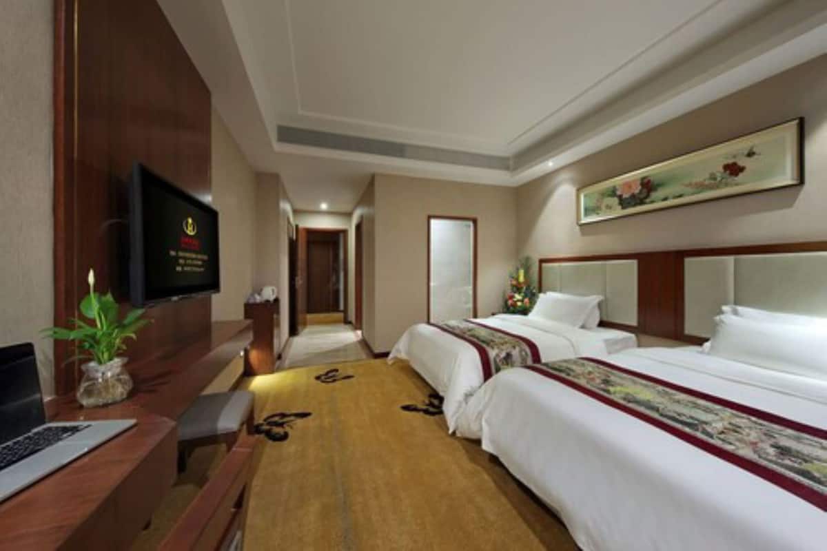 Quarto de hotel com duas camas, uma mesa com um laptop, uma televisão e um quadro na parede.