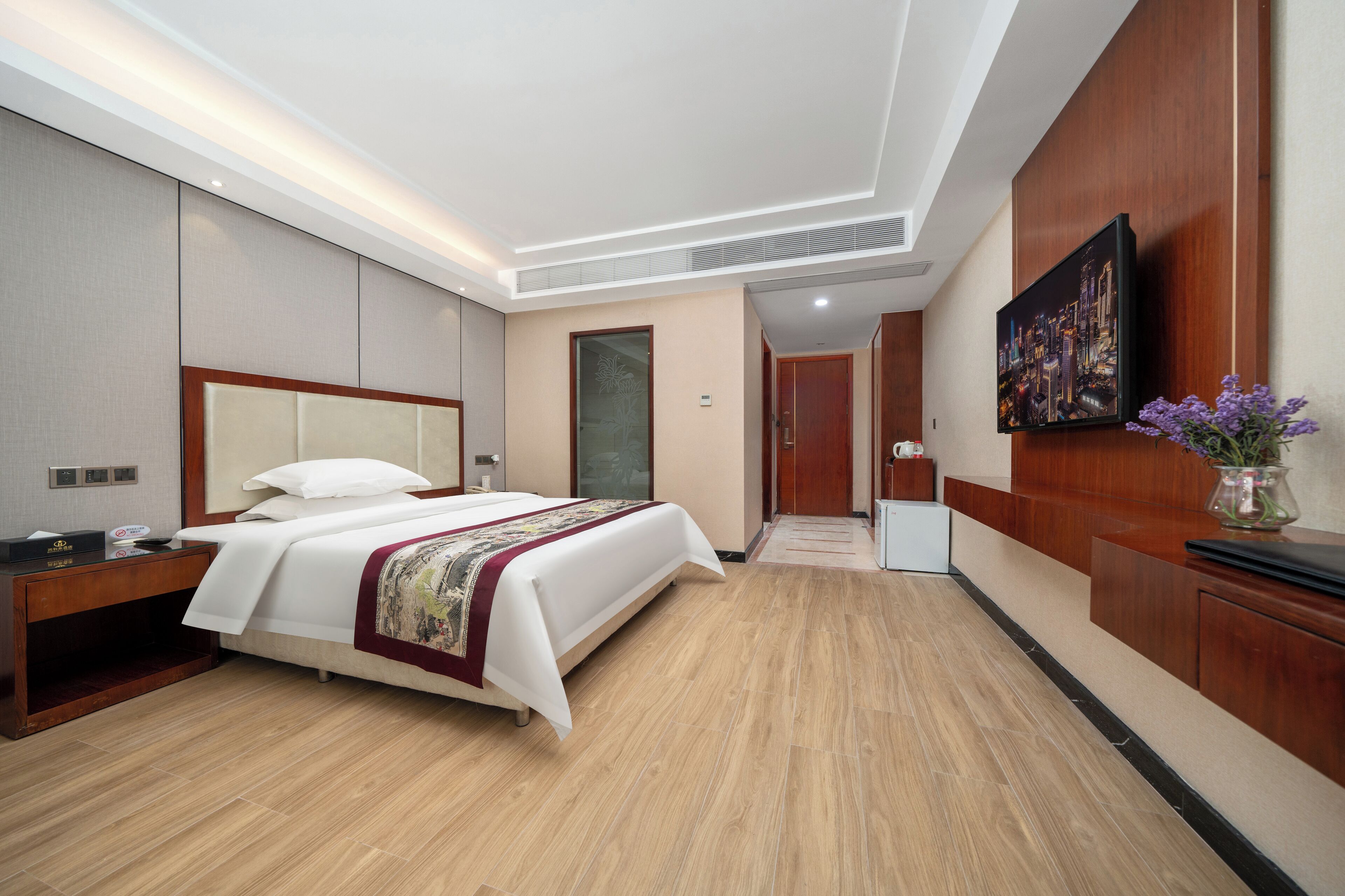 Habitación de hotel con una cama grande, una mesita de noche de madera, un escritorio, una silla, un televisor y un florero con flores.