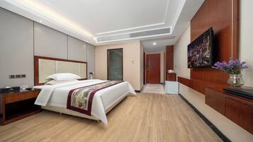 Habitación de hotel con una cama grande, una mesita de noche de madera, un escritorio, una silla, un televisor y un florero con flores.