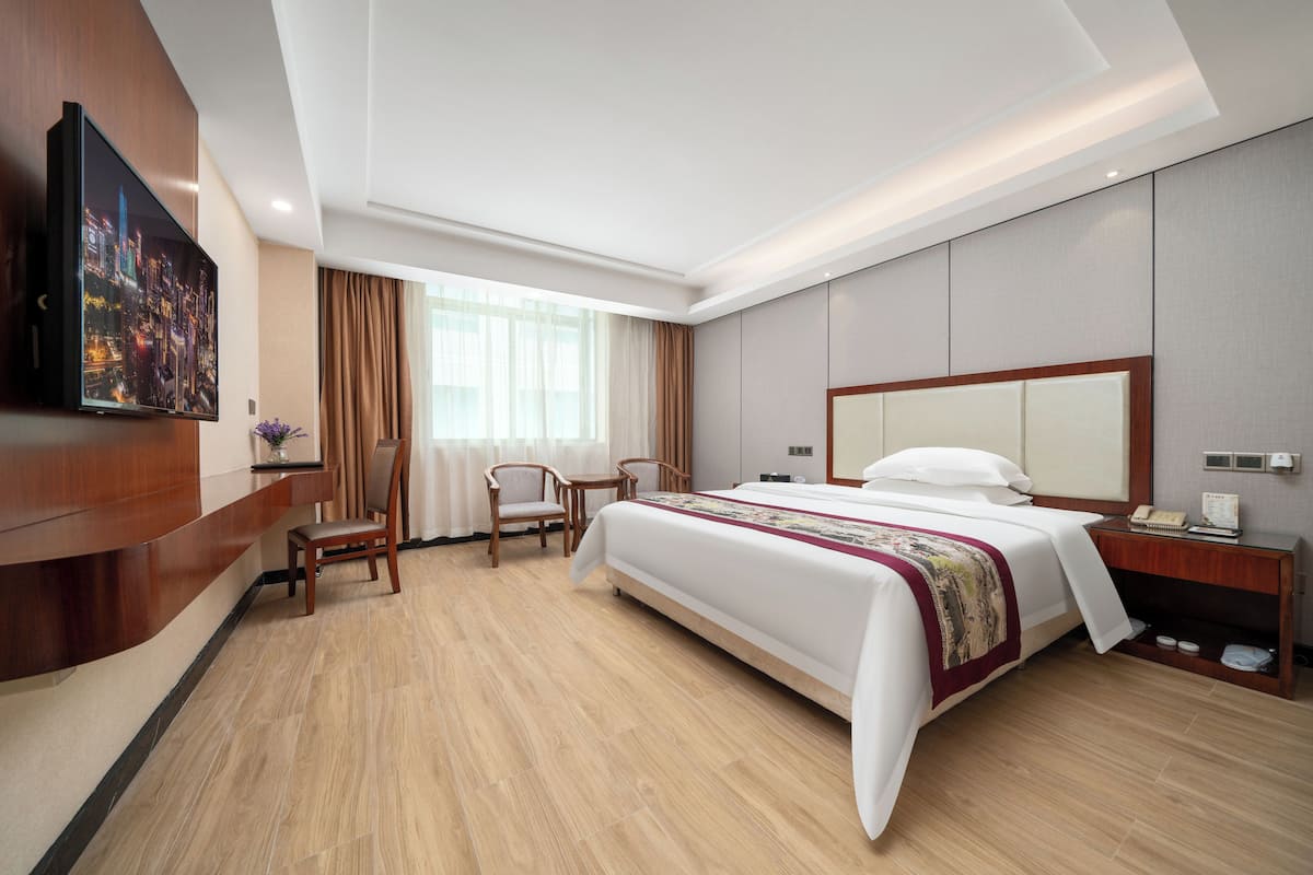Quarto de hotel com uma cama grande, uma escrivaninha com cadeira, uma televisão na parede e uma janela com cortinas.