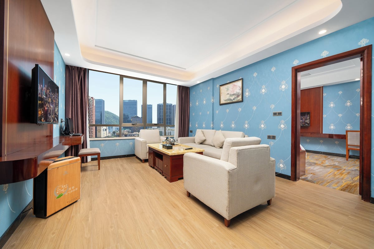 Uma sala de estar moderna com um sofá, uma mesa de centro e uma televisão. Há uma janela grande com vista para a cidade, e as paredes são azuis com um padrão floral.