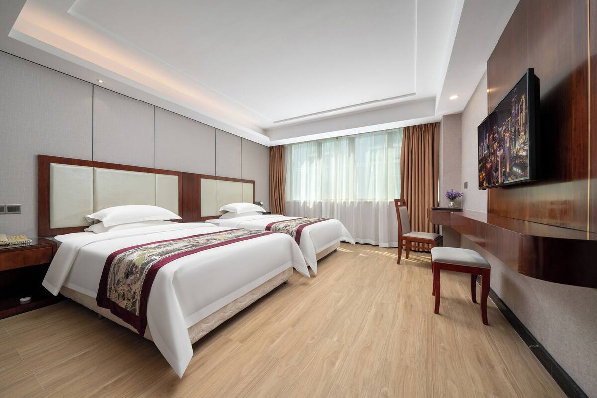 Quarto de hotel com duas camas, piso de madeira, televisão na parede e janela com cortinas.