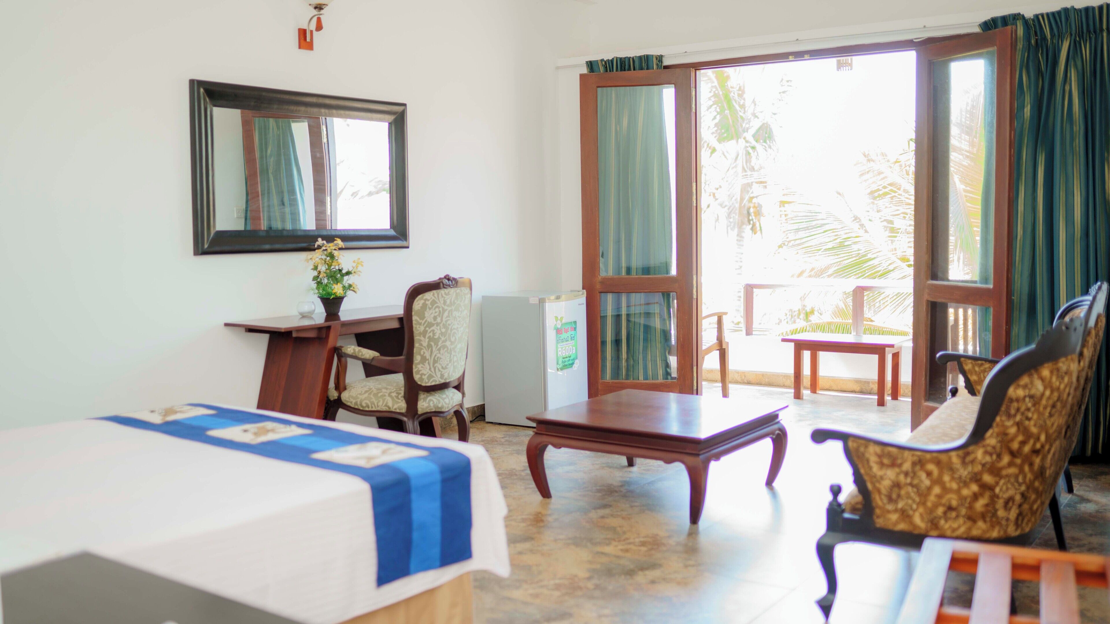 Habitación de hotel con cama, escritorio, silla, mesita pequeña y balcón con vista a palmeras.