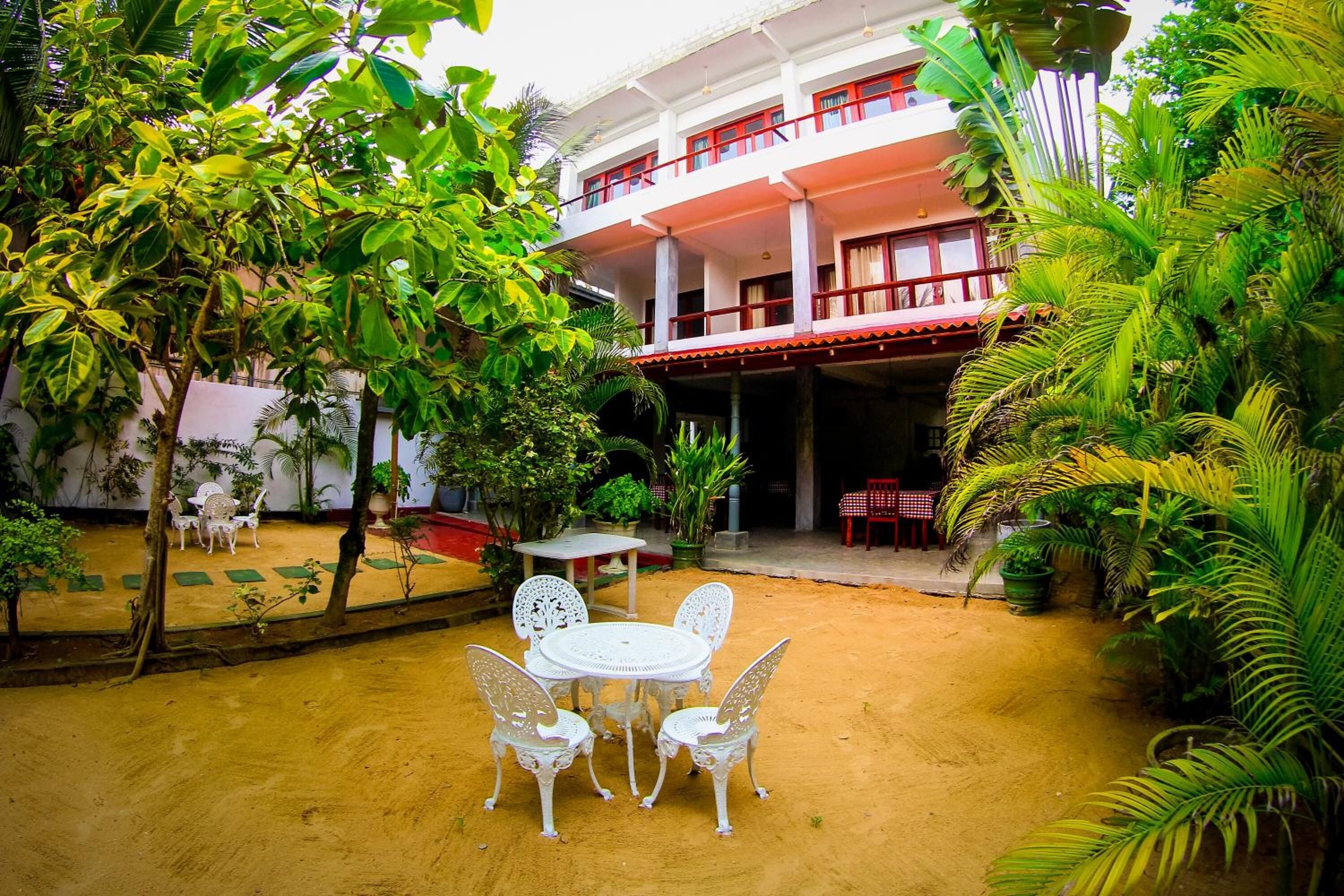 Um pátio com uma mesa e cadeiras brancas, rodeado por vegetação exuberante e um edifício com várias varandas.