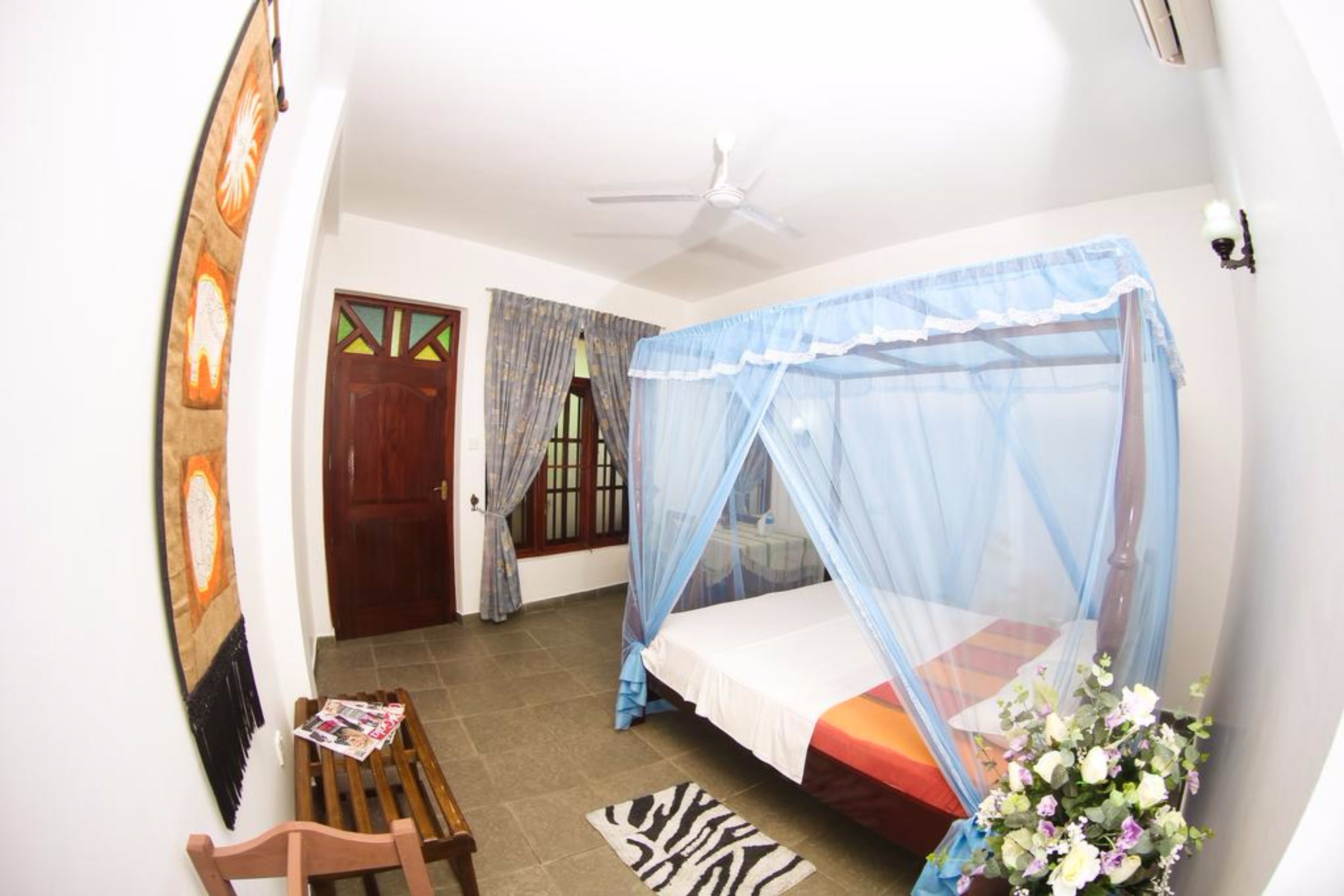 A bedroom with a bed under a mosquito net, a wooden chair, a zebra-patterned rug, and a wall with a framed picture.