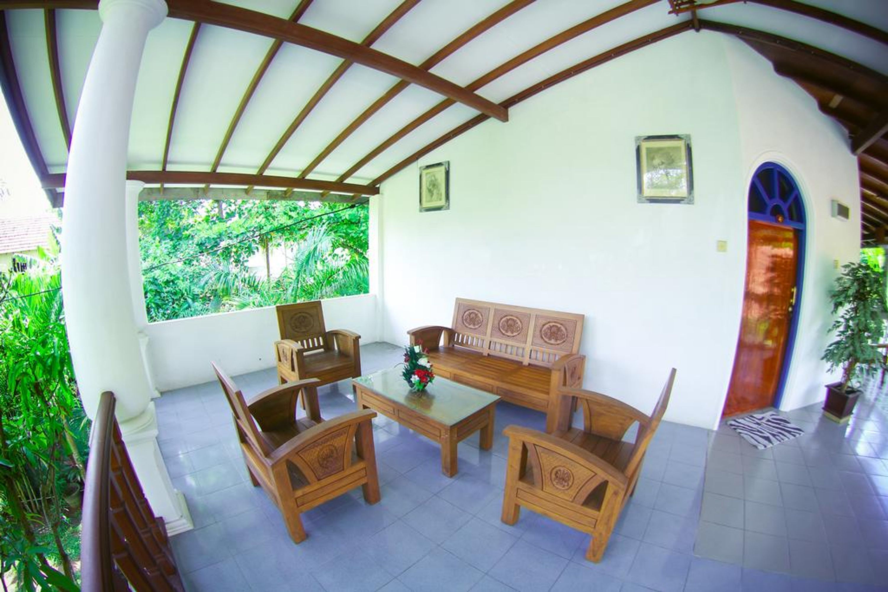A covered outdoor seating area with wooden furniture and a glass table, surrounded by white walls and a view of greenery.