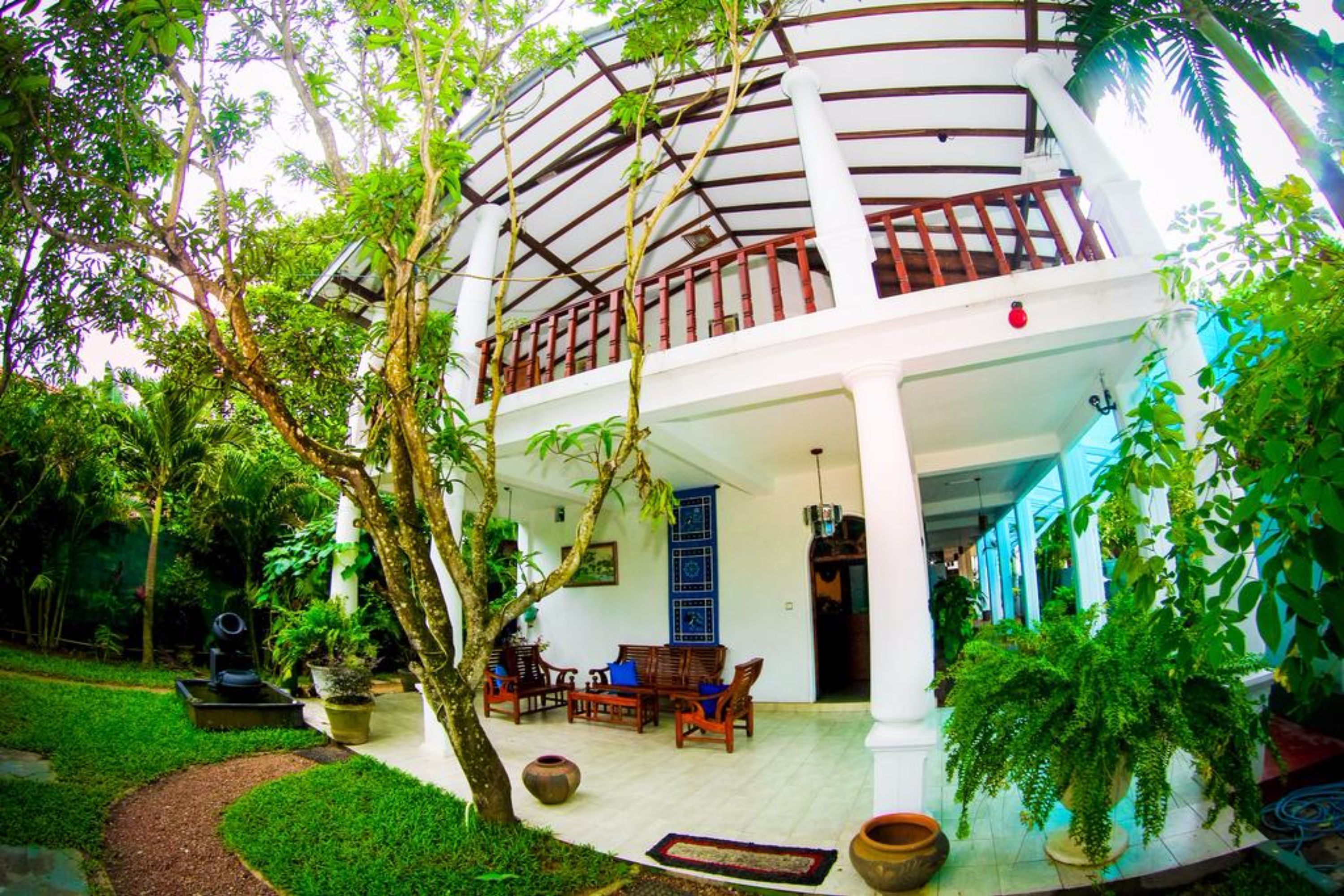 A two-story building with a covered balcony, surrounded by lush greenery and potted plants.