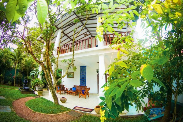 A two-story building with a covered balcony, surrounded by lush greenery and potted plants.