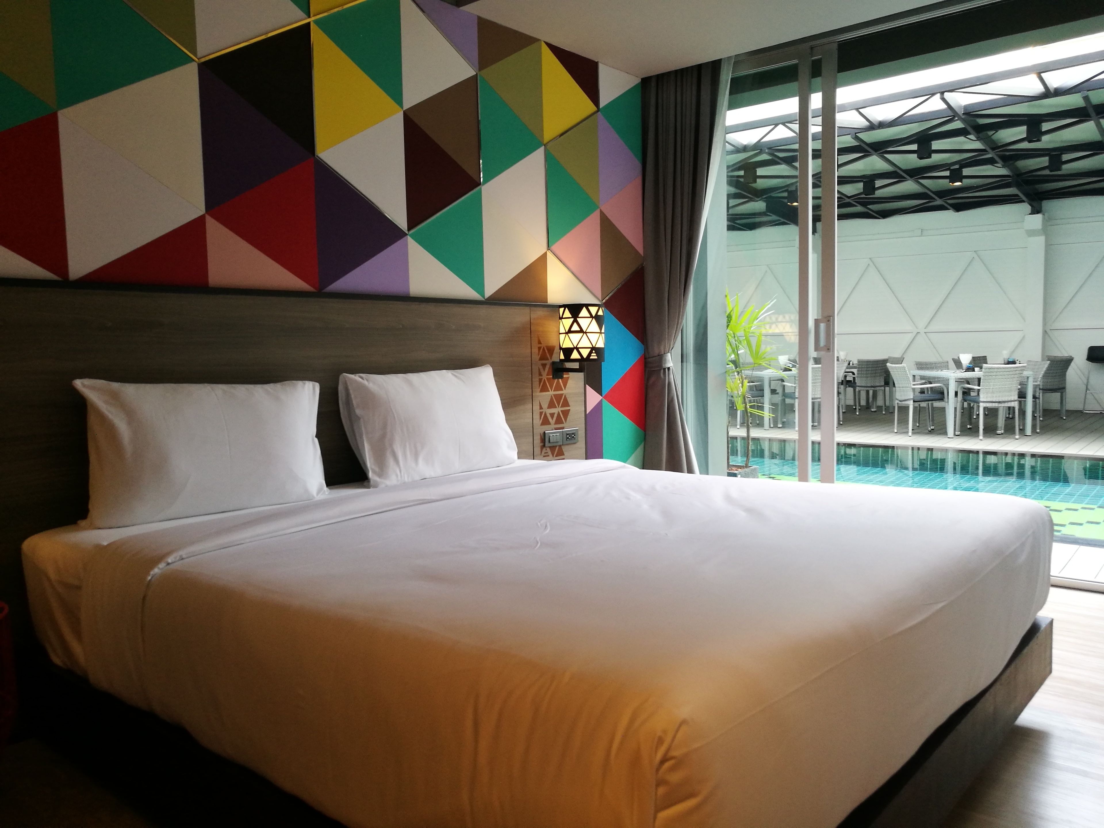 A hotel room with a large bed, a colorful geometric wall, a nightstand with a lamp, and a view of an outdoor pool area.