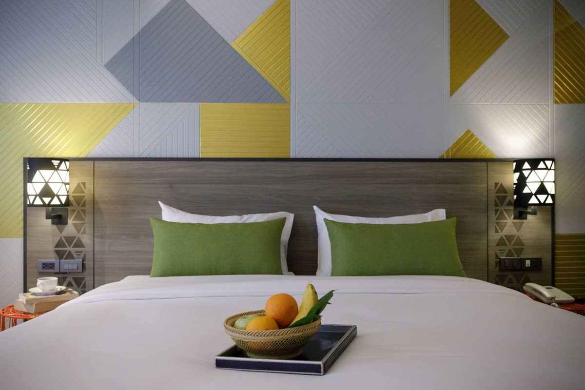 A hotel room with a large bed, two green pillows, a bedside table with a phone, and a wall with geometric patterns.