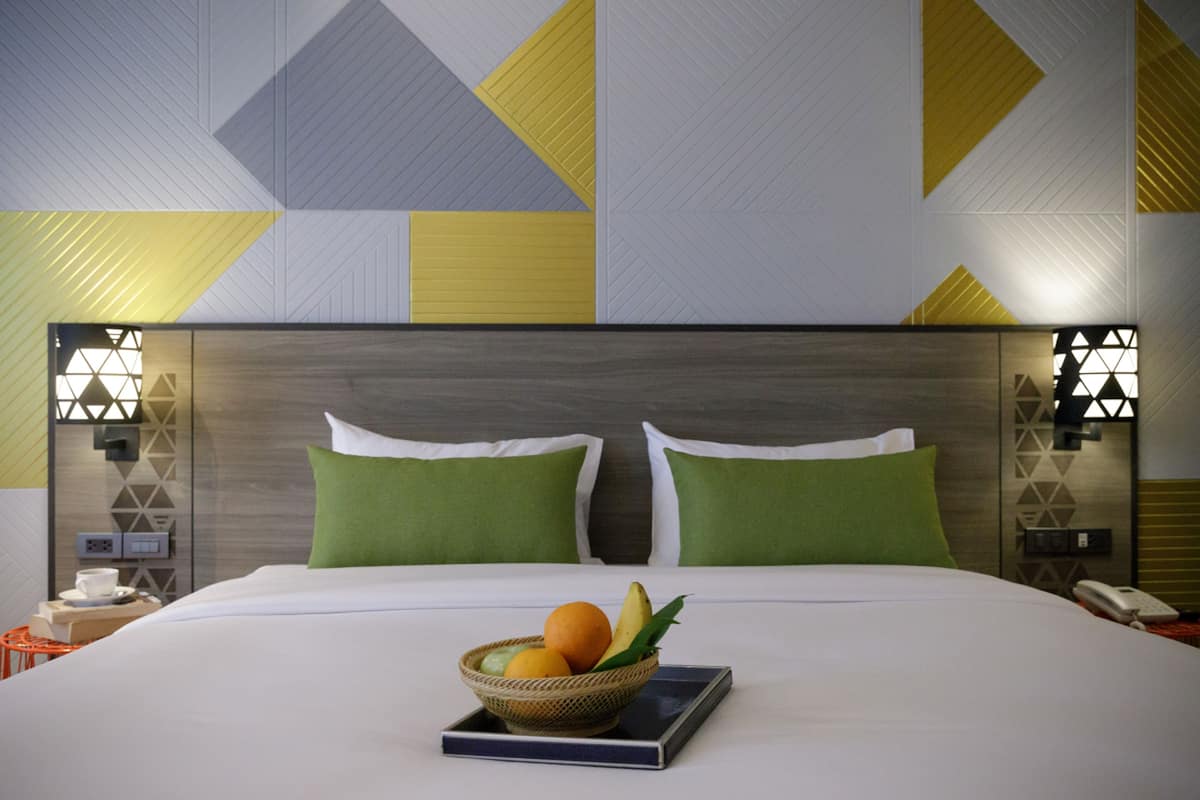 A hotel room with a large bed, green pillows, a bedside table with a phone, and a wall with geometric patterns.