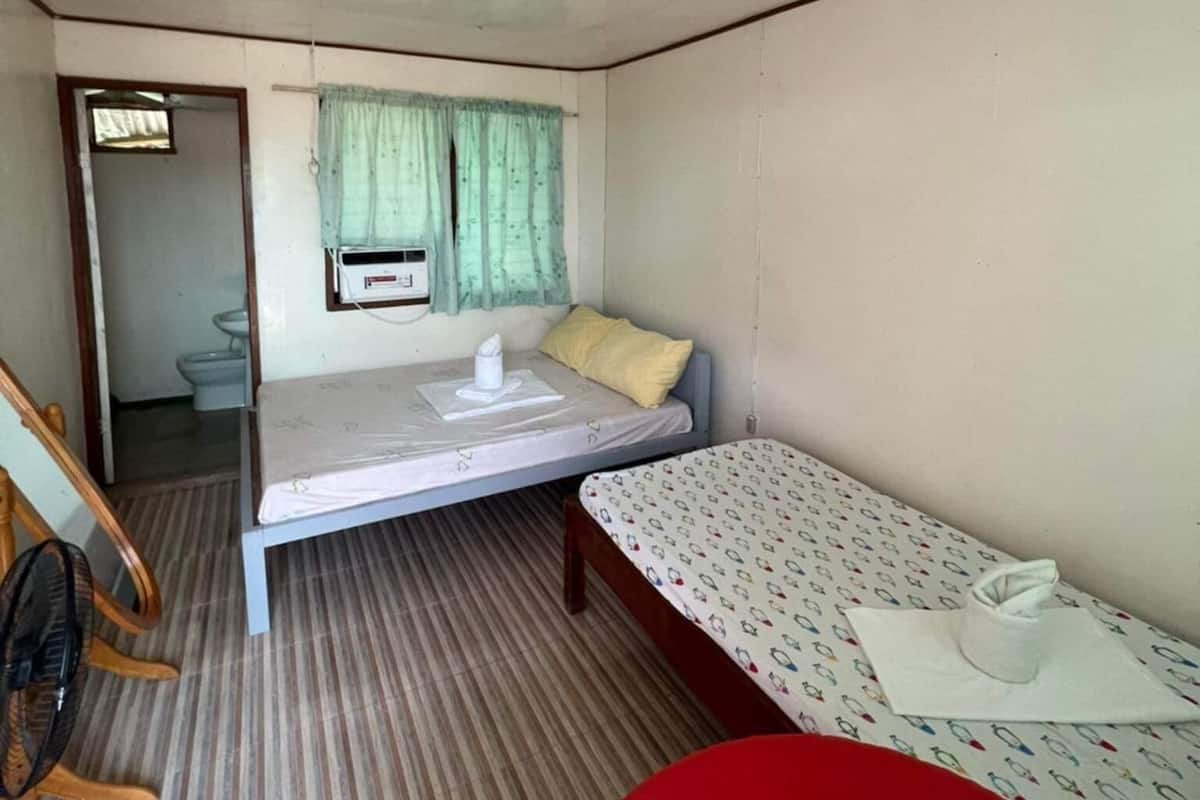 A small, compact room with a single bed, a small table, and a chair. There is a window with a curtain, a light fixture on the ceiling, and a door leading to another room.