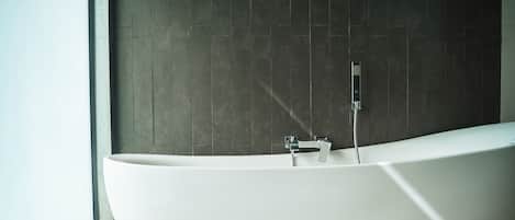 A modern, freestanding bathtub in a bathroom with dark tiled walls and a window to the left.