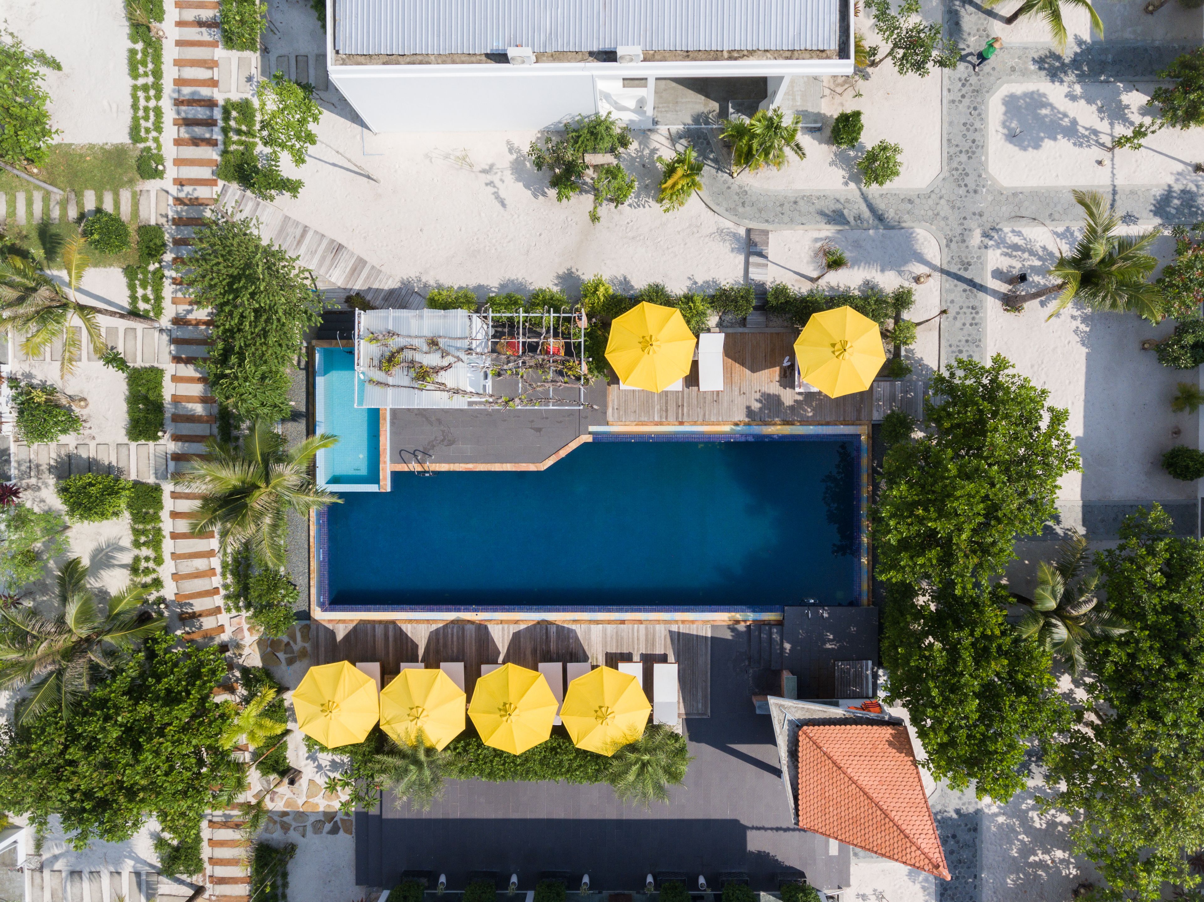 Vue aérienne d’un complexe hôtelier doté d’une piscine centrale, entourée de palmiers et de parasols.