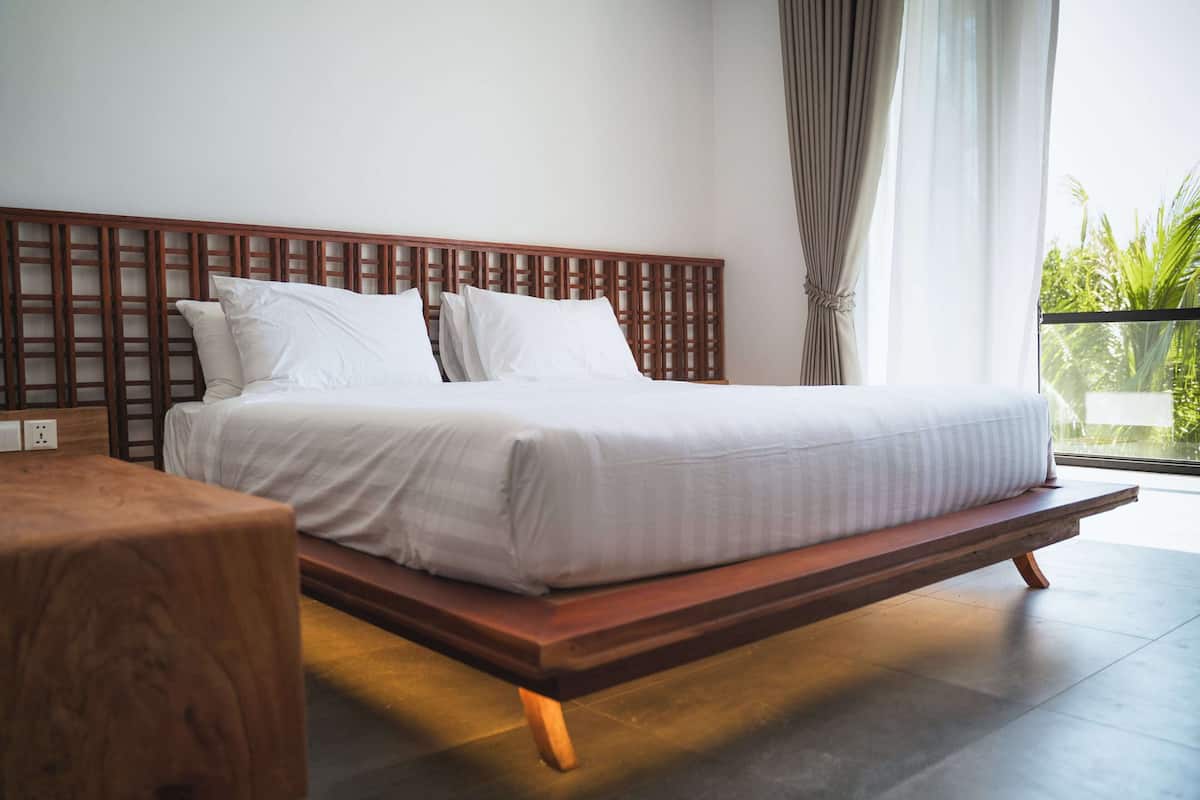 A modern bedroom with a wooden headboard, a wooden bedside table, and a large window with curtains.