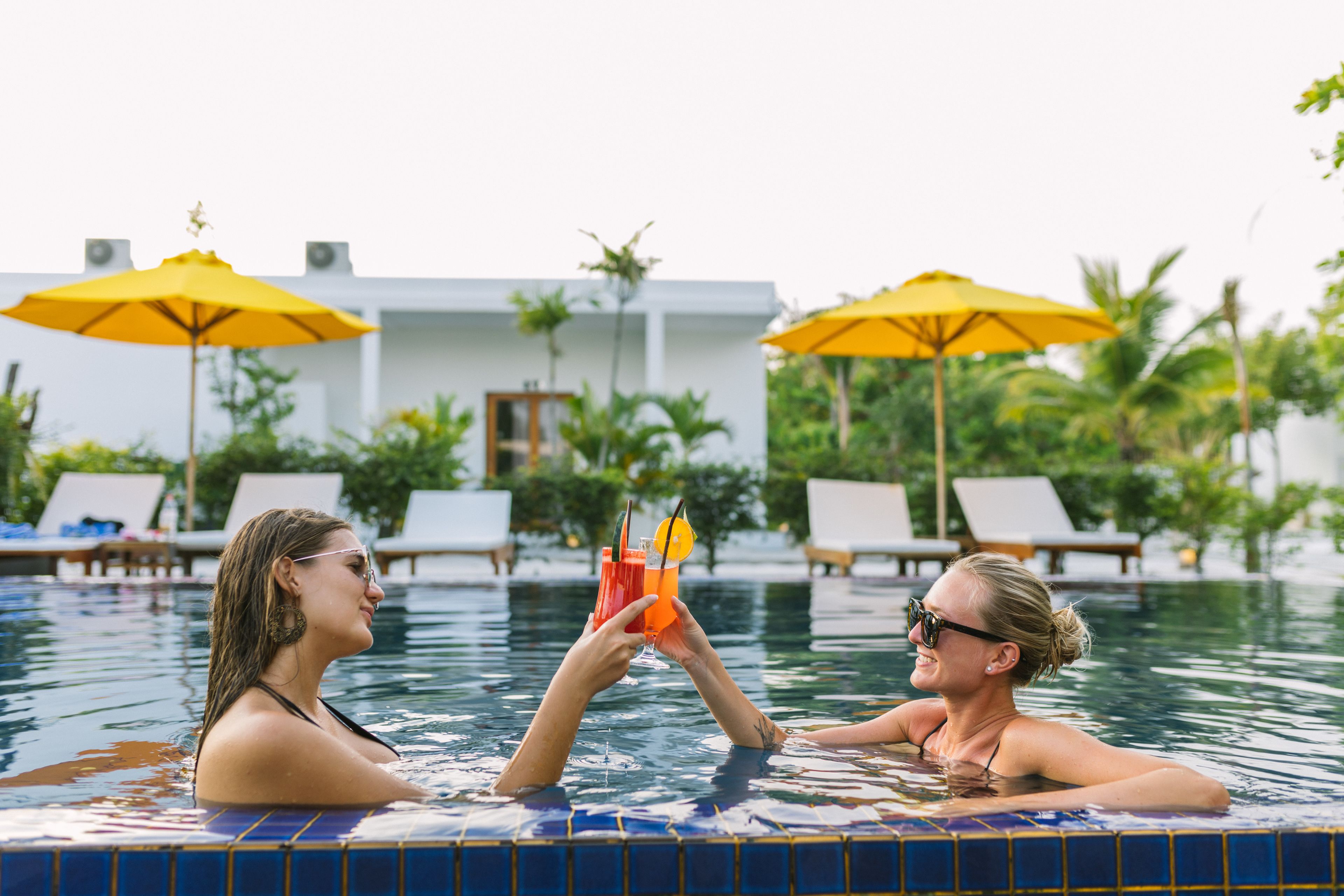 A swimming pool with two people toasting with drinks, surrounded by lounge chairs and yellow umbrellas.