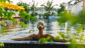 Eine Person in einem Schwimmbecken mit Fliesenrand, umgeben von Grünpflanzen und einem Gebäude im Hintergrund.