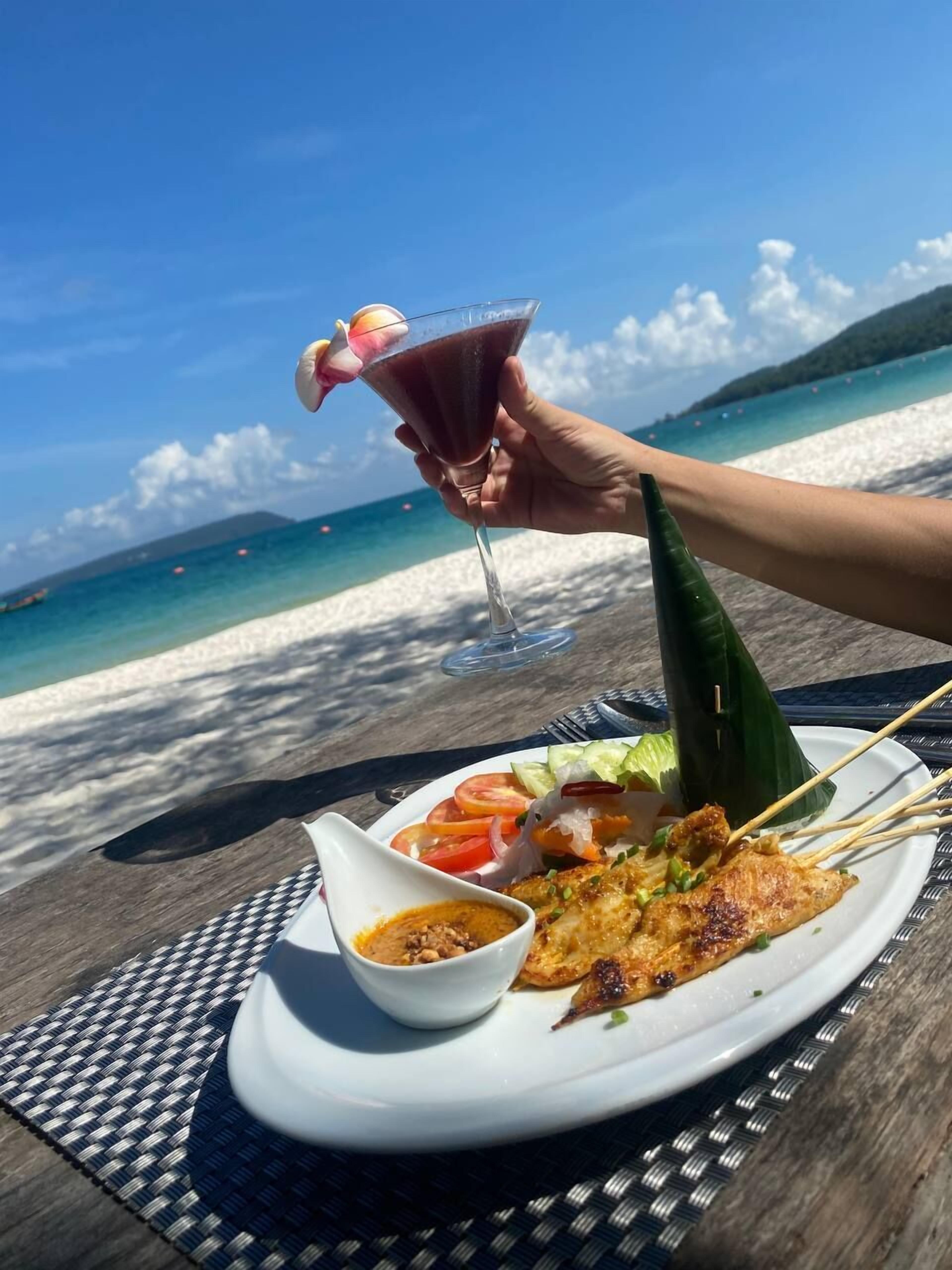 A meal with grilled chicken, vegetables, and a drink on a table by the beach.