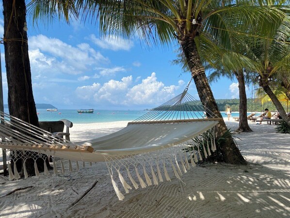 A hammock on a sandy beach with palm trees, a clear blue sky, and a calm sea in the background.