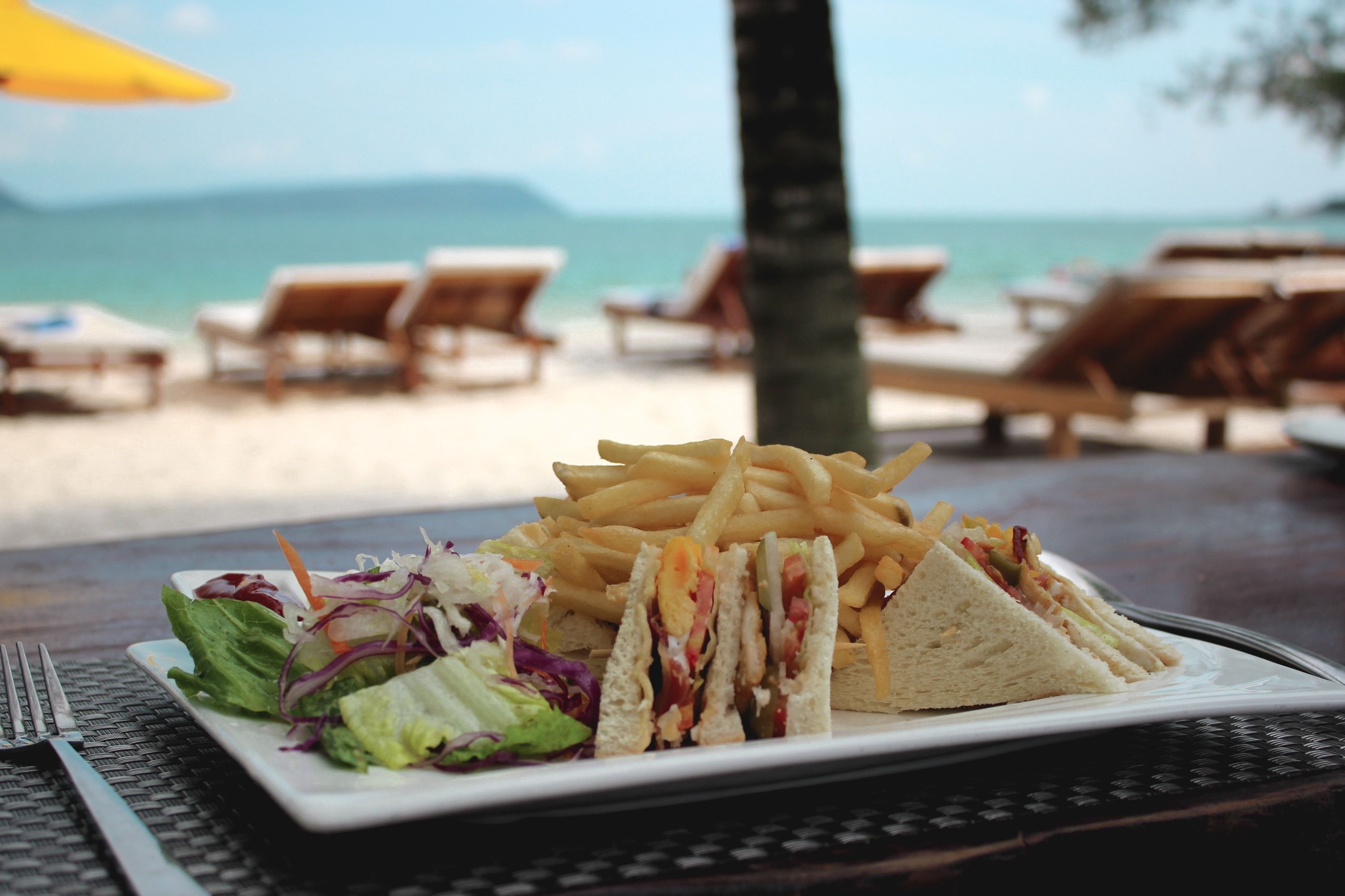 A meal of a sandwich, fries, and salad on a table with a view of the beach and ocean in the background.
