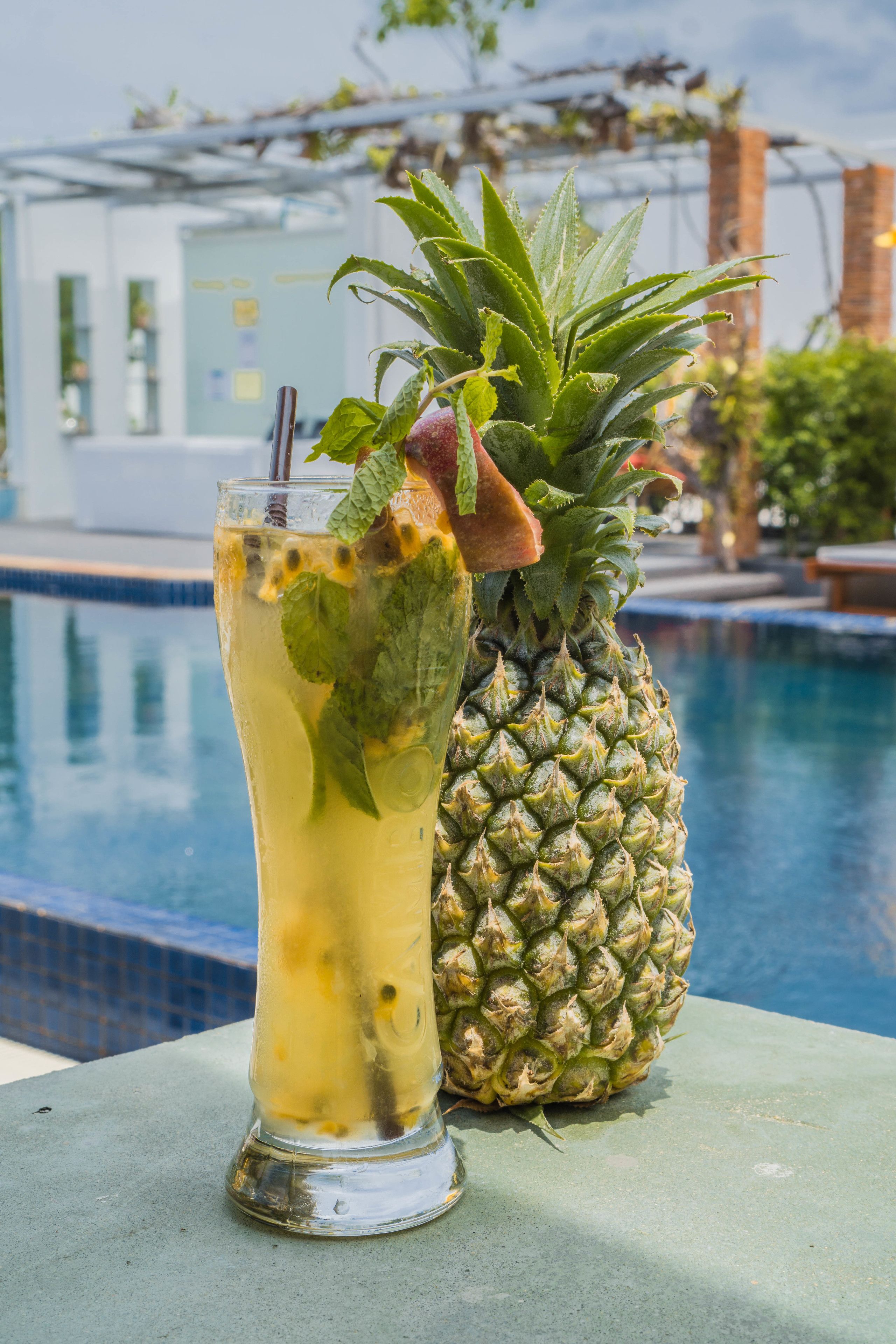 A glass of a yellow beverage with mint and a pineapple beside a pool.
