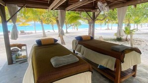 Zwei hölzerne Betten mit weißen Leinen und Kissen unter einer Holzkonstruktion, mit Blick auf einen Strand mit Palmen und das Meer.