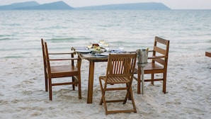 Ein Holztisch mit Tellern, Gläsern und einem Eimer auf einem sandigen Strand, mit Stühlen, Blick auf das Meer und entfernte Berge.