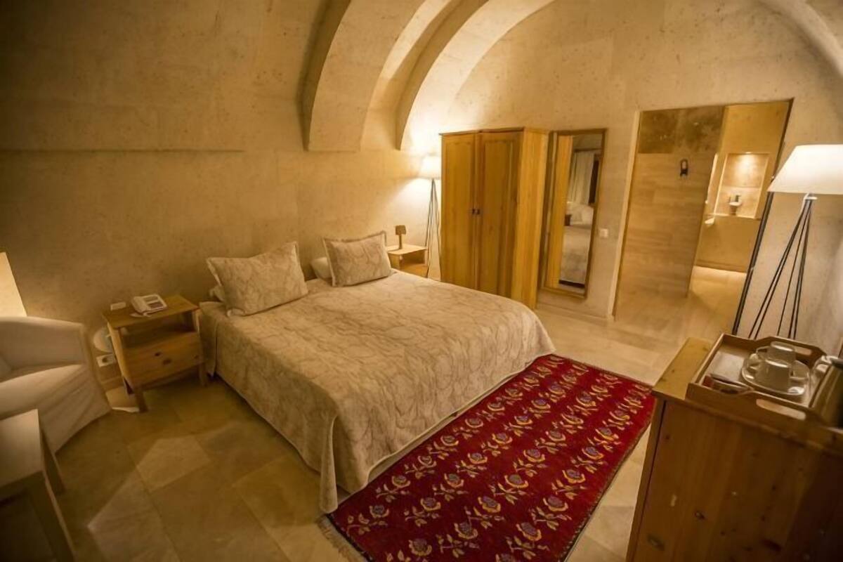 A bedroom with a large bed, a red patterned rug, a wooden wardrobe, and a nightstand with a lamp.