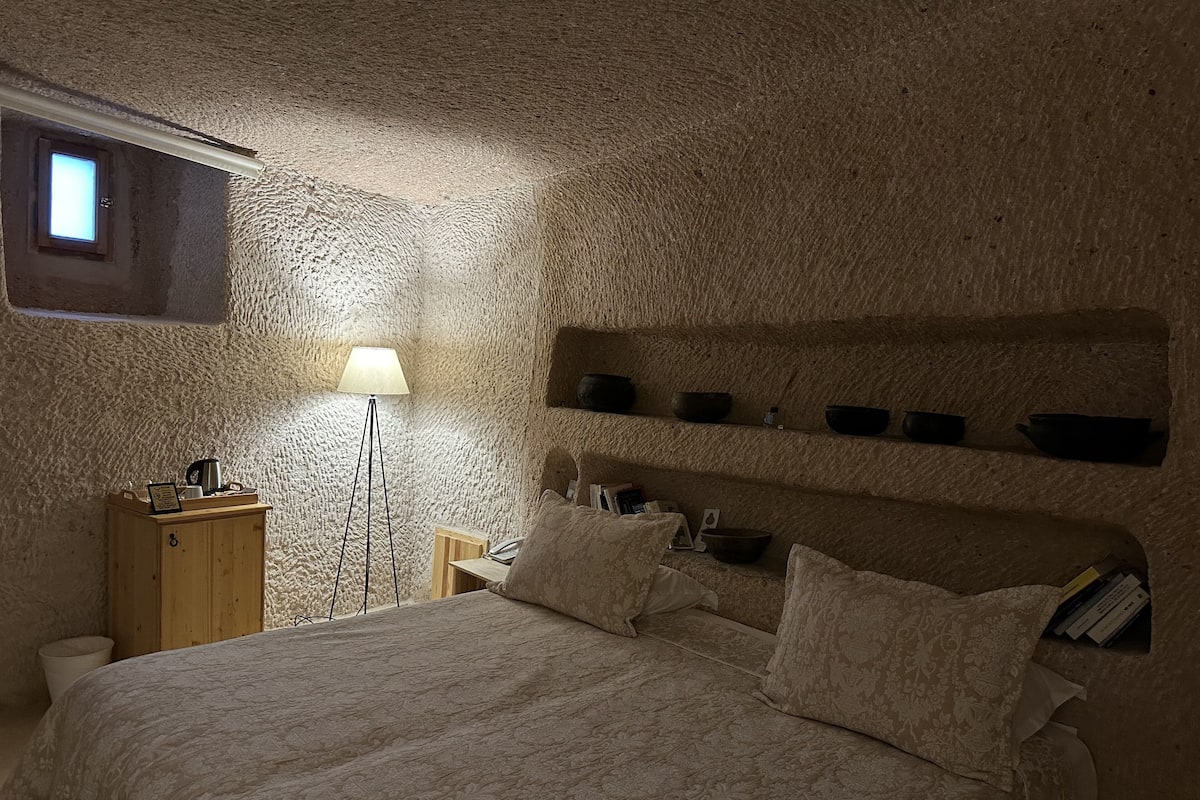 A bedroom with a textured stone wall, a bed with patterned bedding, a bedside table with a lamp, and a shelf with bowls.