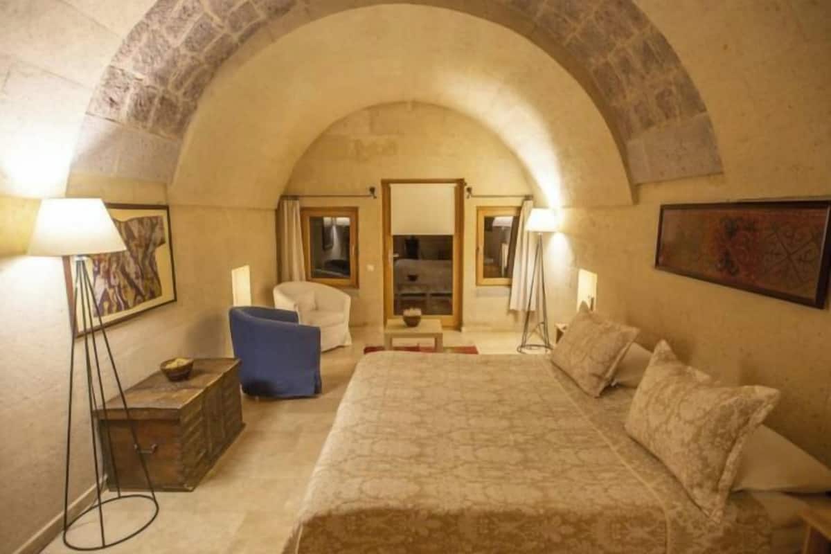 A bedroom with a large bed, a blue chair, a wooden chest, and a stone archway.