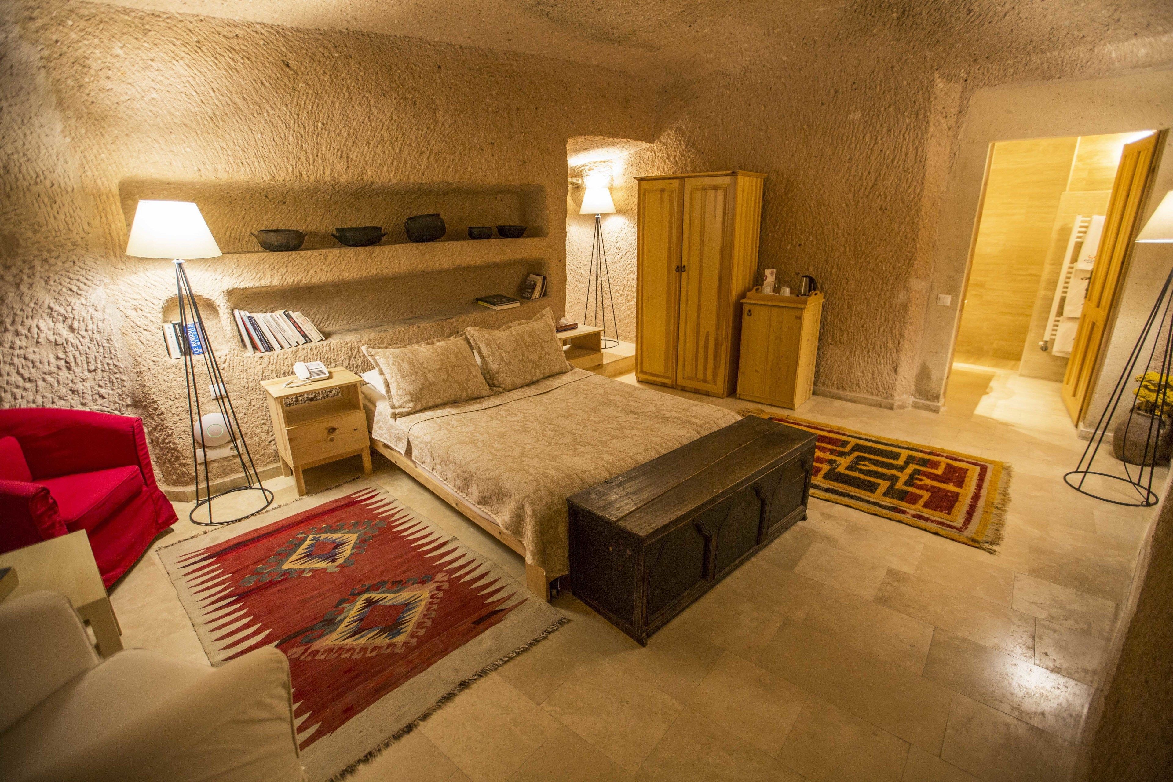 Un dormitorio con cama, mesitas de noche, un armario, un sillón rojo, una lámpara de pie y una alfombra estampada.