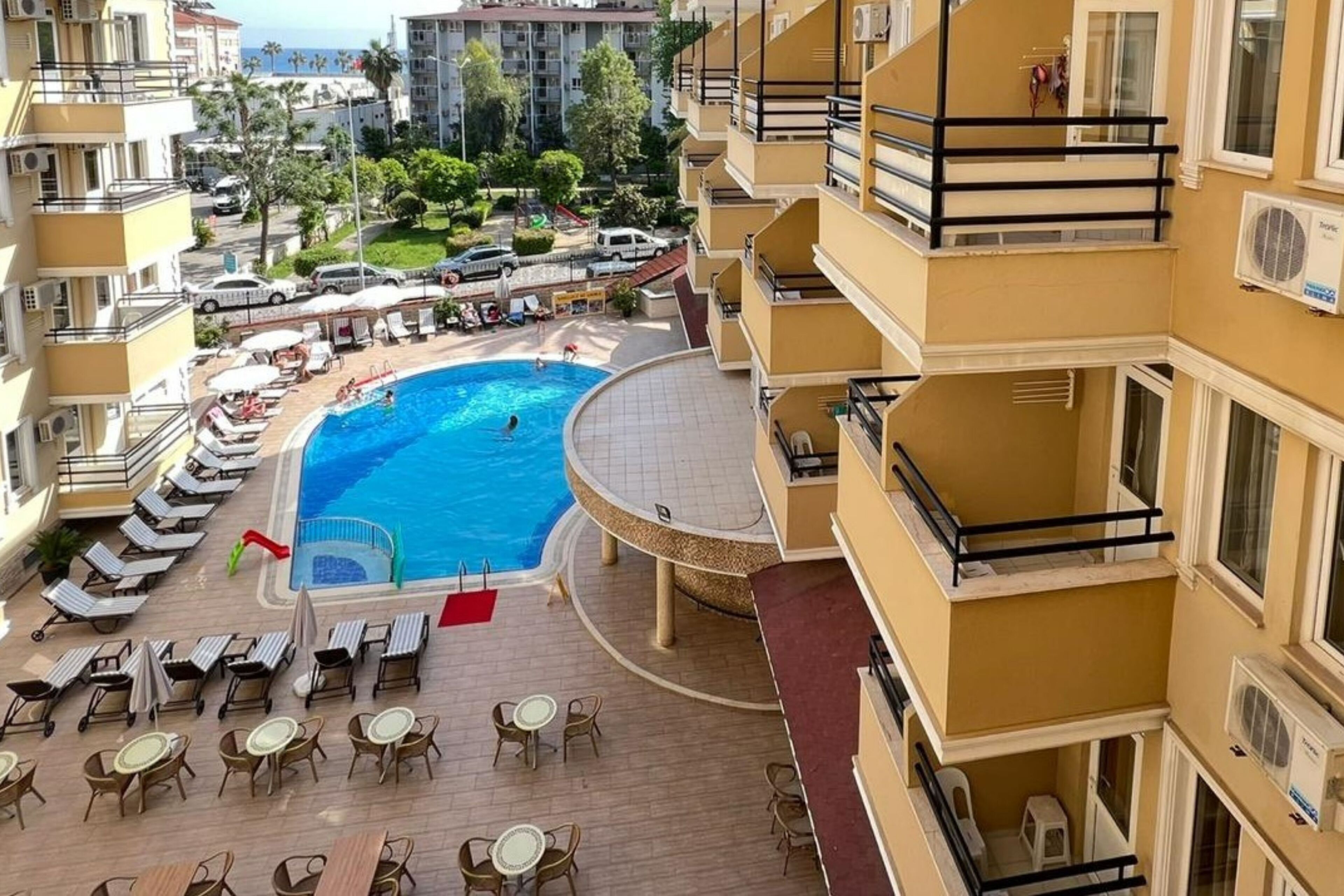 A rooftop pool area with lounge chairs and tables, surrounded by a yellow building with balconies.