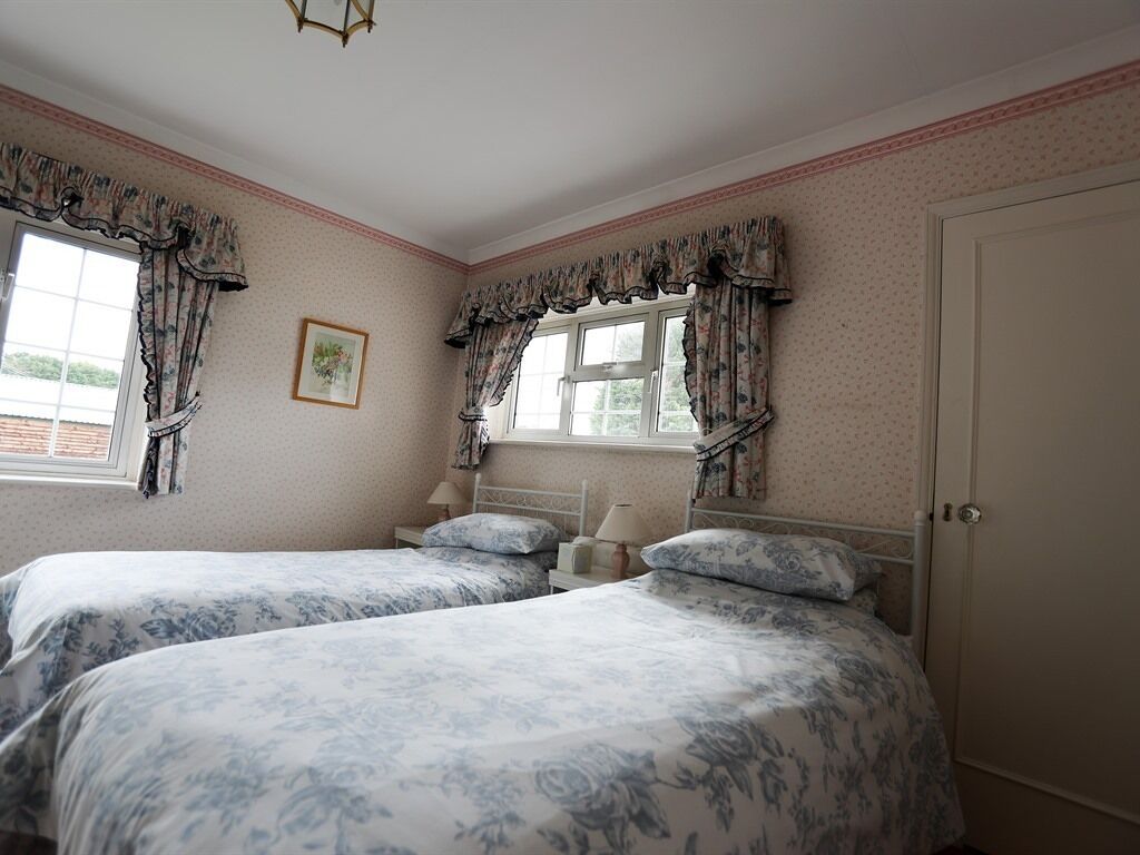 A bedroom with two beds, floral-patterned bedding, a window with curtains, a bedside table with a lamp, and a door.