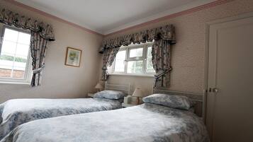 A bedroom with two beds, floral-patterned bedding, a window with curtains, a bedside table with a lamp, and a door.