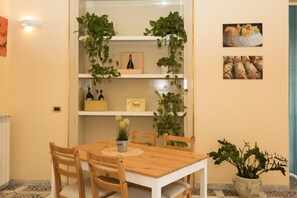 Un comedor con mesa y sillas de madera, rodeado de macetas con plantas y estantes con botellas de vino y un cuadro enmarcado.