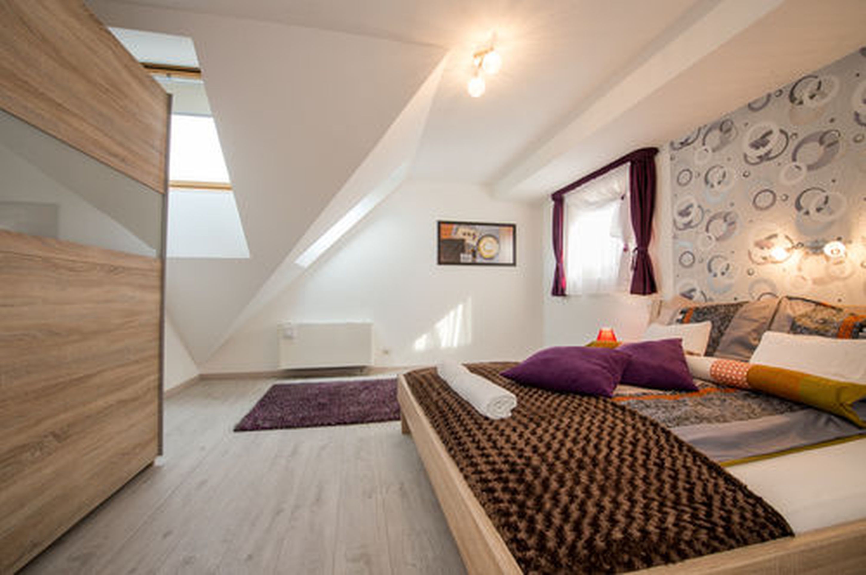 A bedroom with a sloped ceiling, a bed with patterned bedding, a window with curtains, and a wall-mounted light fixture.