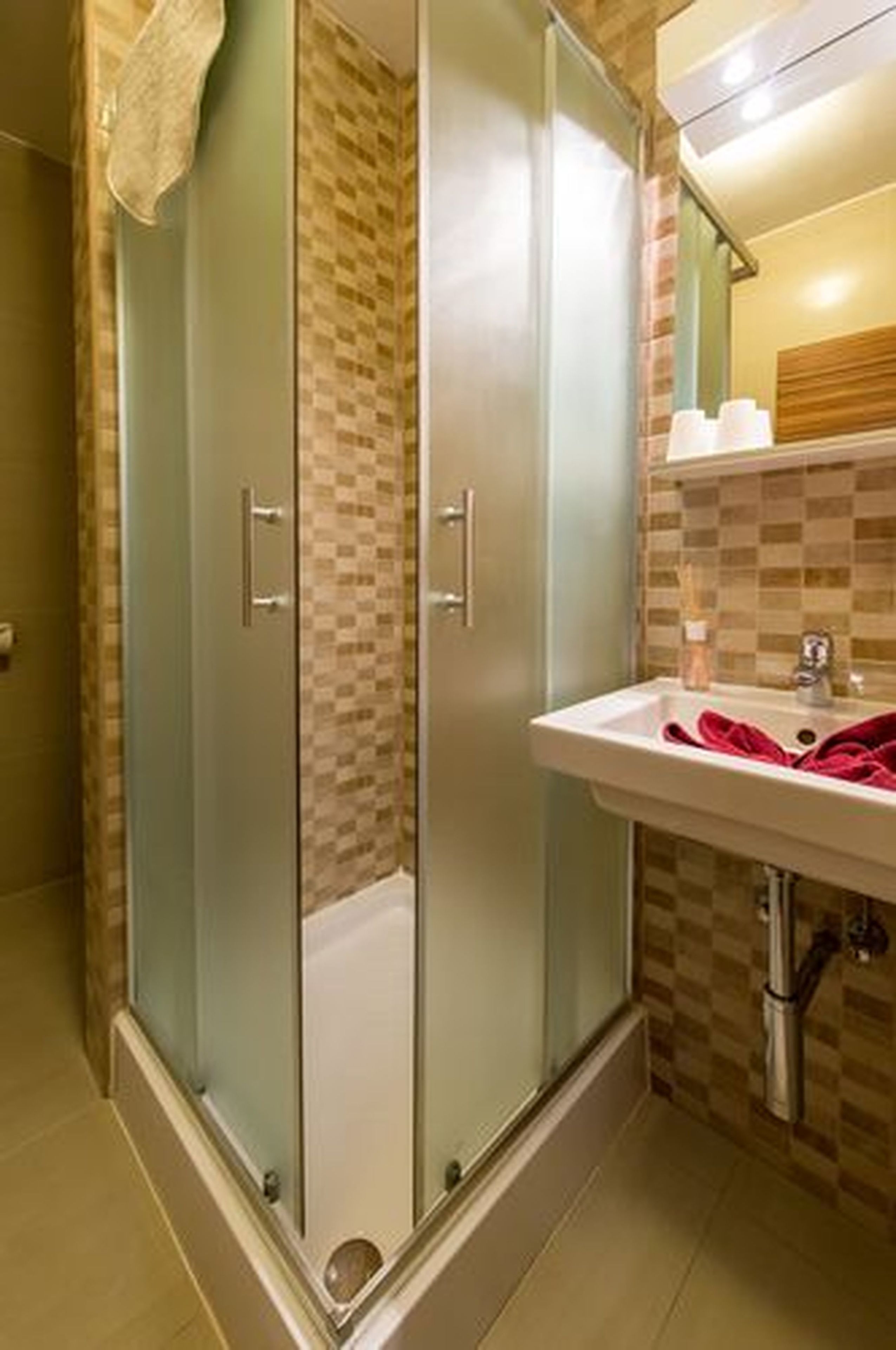 Une salle de bain avec une douche à parois de verre, un lavabo surmonté d’une serviette rouge et un mur recouvert de carreaux.