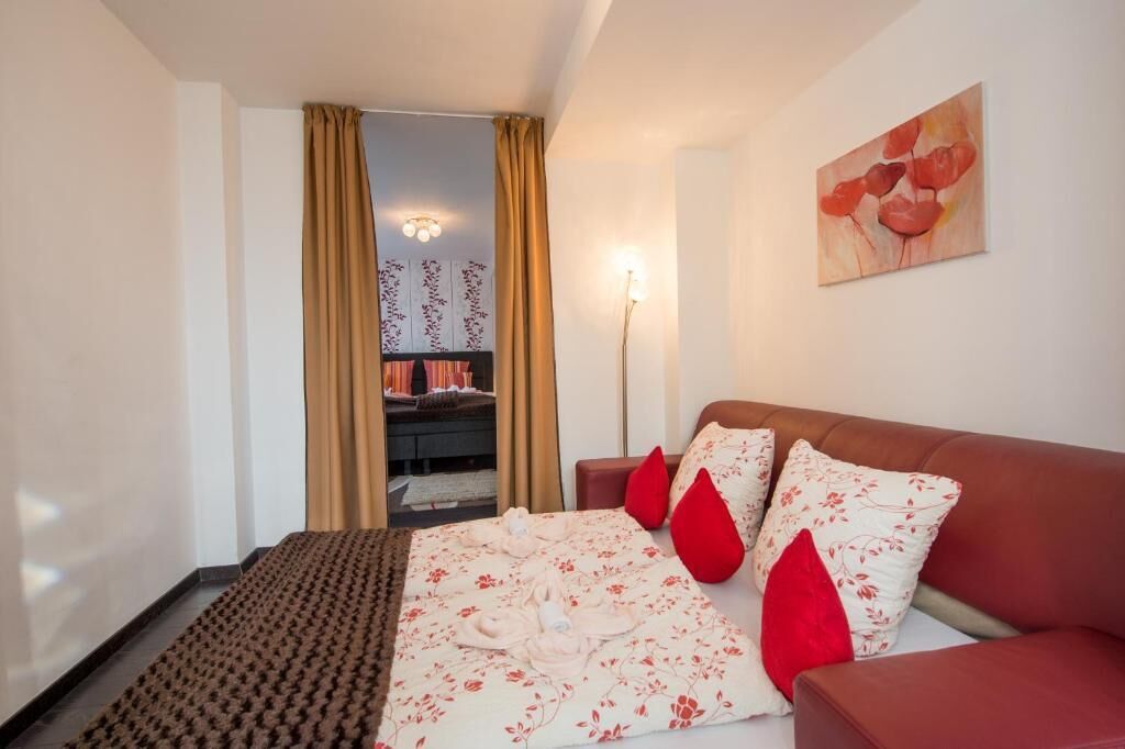 Ein Hotelzimmer mit einem Bett, einem roten Sofa, einem Gemälde an der Wand und Blick in einen anderen Raum durch einen Vorhang.