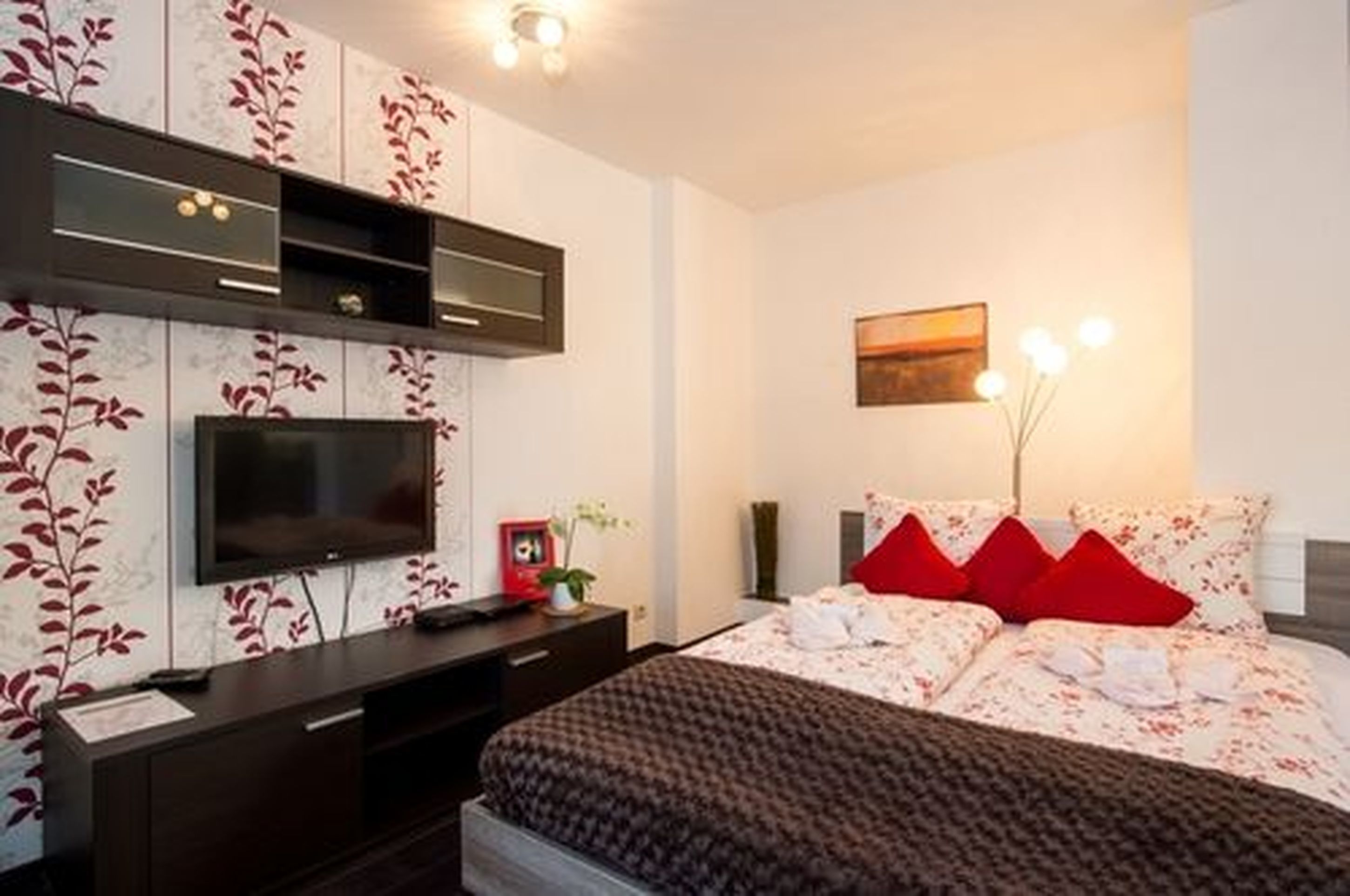 A modern bedroom with a flat-screen TV, a bedside table, a bed with red pillows, and a wall-mounted shelf.