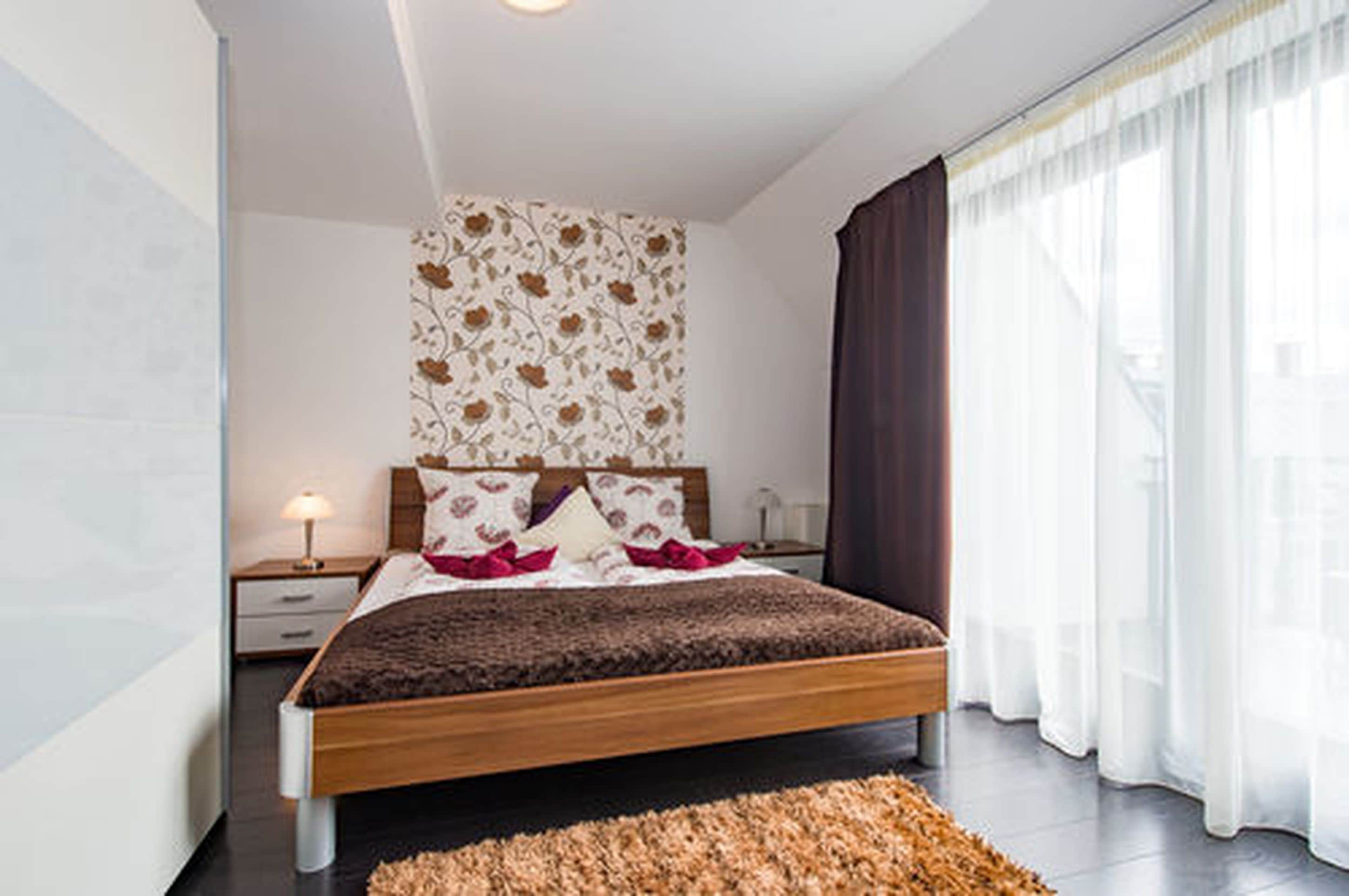 A modern bedroom with a wooden bed, a patterned wall, and a large window with curtains.