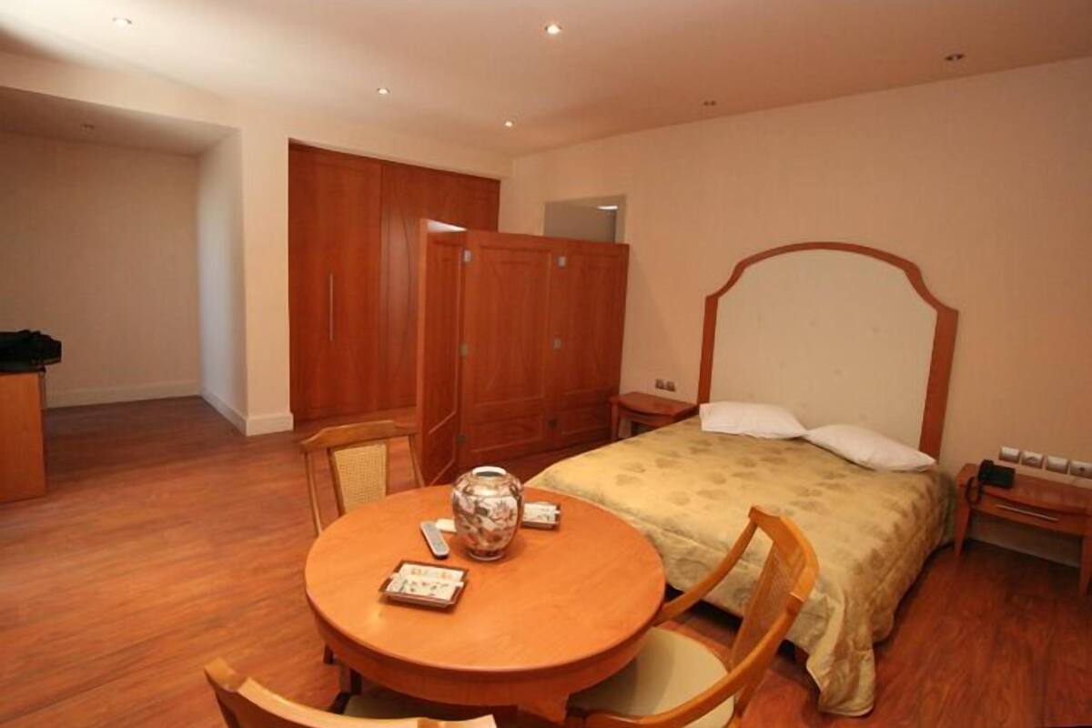 A bedroom with a wooden bed, a round wooden table with chairs, a wardrobe, and a bedside table with a lamp.