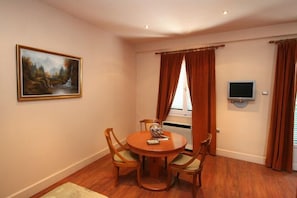 A dining area with a round table and four chairs, a painting on the wall, a television mounted on the wall, and a window with curtains.