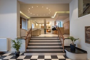 A modern interior with a checkered floor, a staircase with wooden railings, and potted plants. - Palladion Hotel (Syros)