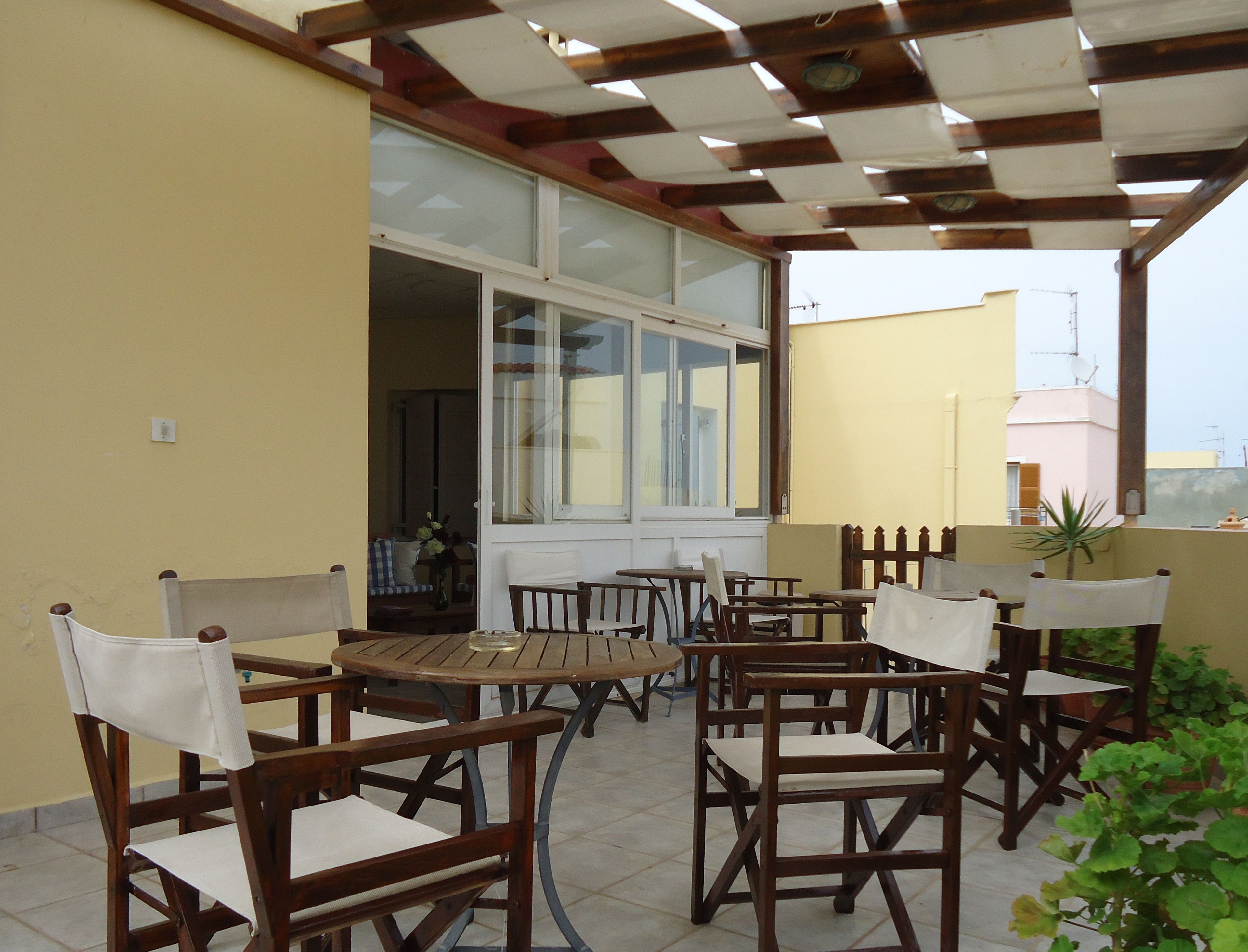 Un patio al aire libre con mesas y sillas de madera, un vidrio que da al interior y vistas del paisaje urbano de fondo.